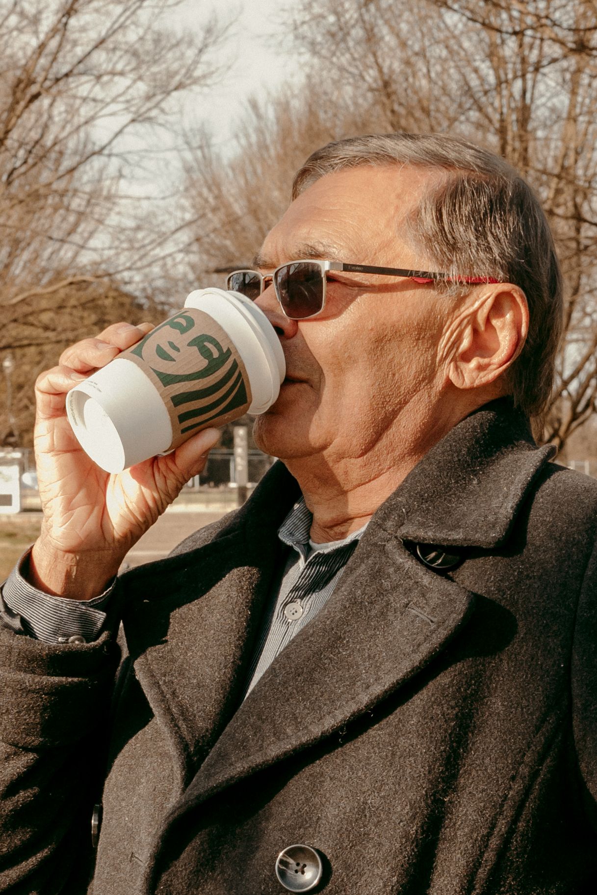 Person in dark coat and sunglasses drinks from a white paper coffee cup outdoors.