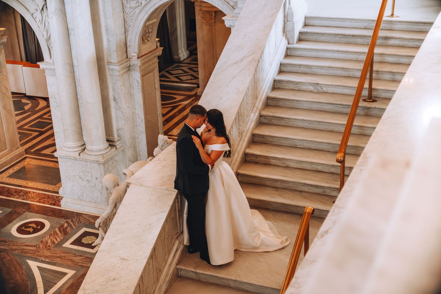 Newlyweds share an intimate moment on a grand marble staircase in an elegant venue.