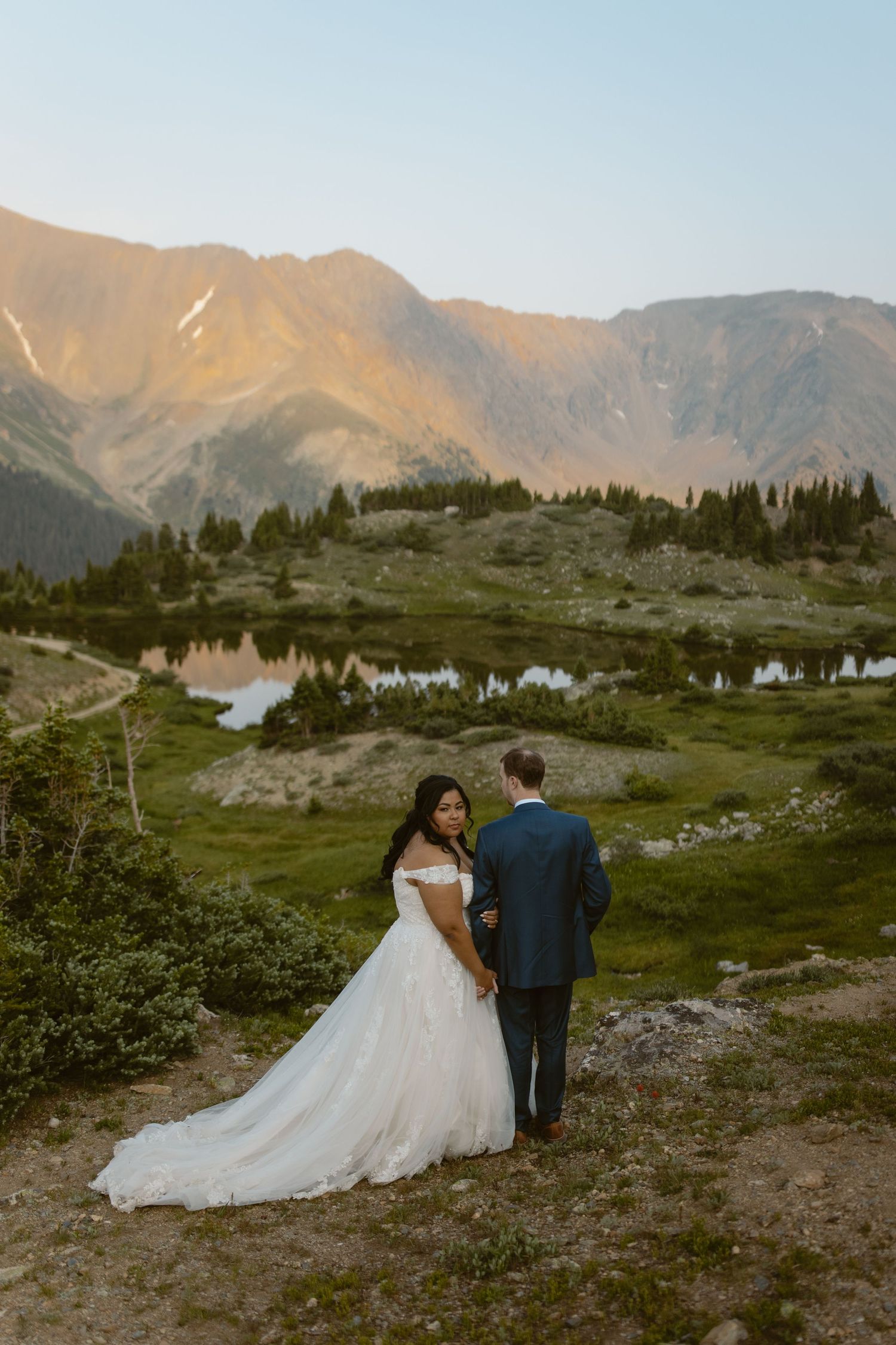 A couple in wedding attire walks along a mountain trail overlooking an alpine lake at sunset in a scenic landscape.