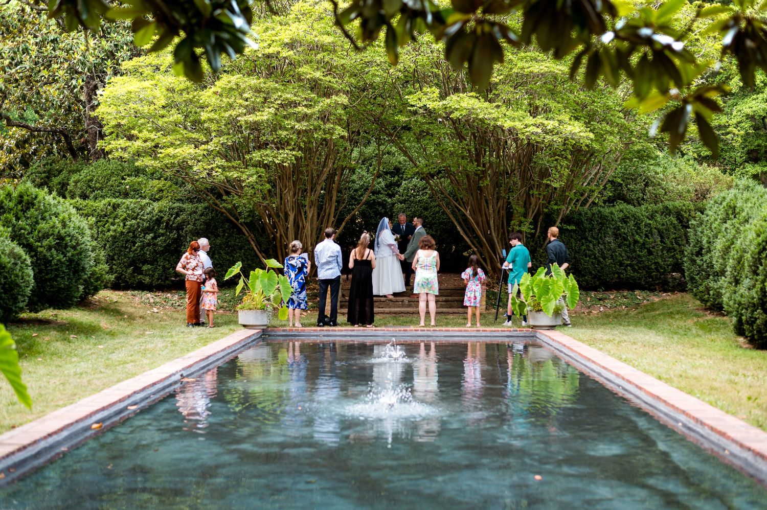 A group follows a garden path between stone walls near a reflective pool surrounded by lush greenery in a formal garden setting.