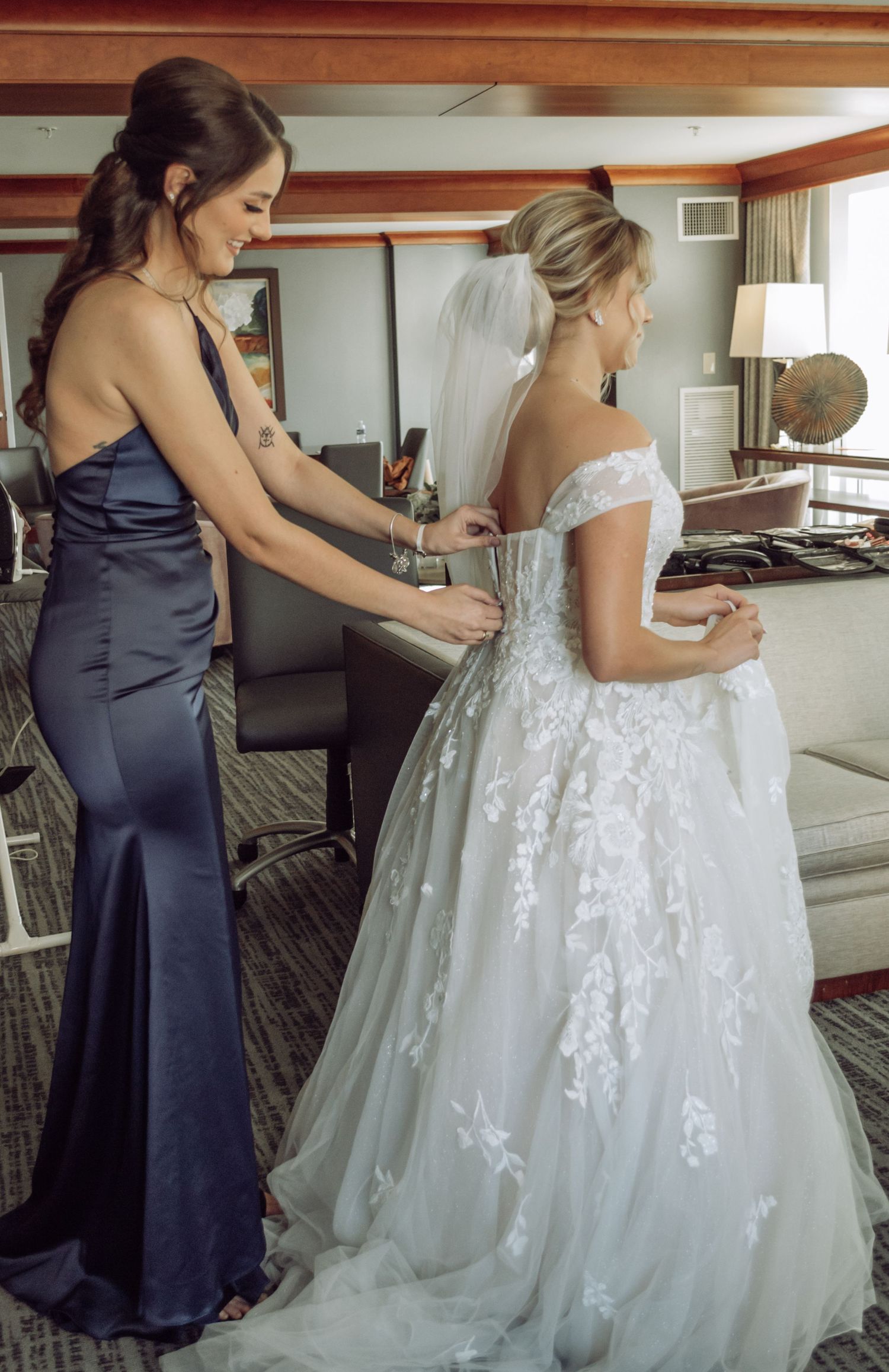 Bridesmaid in navy dress helps adjust white lace wedding gown in hotel room.