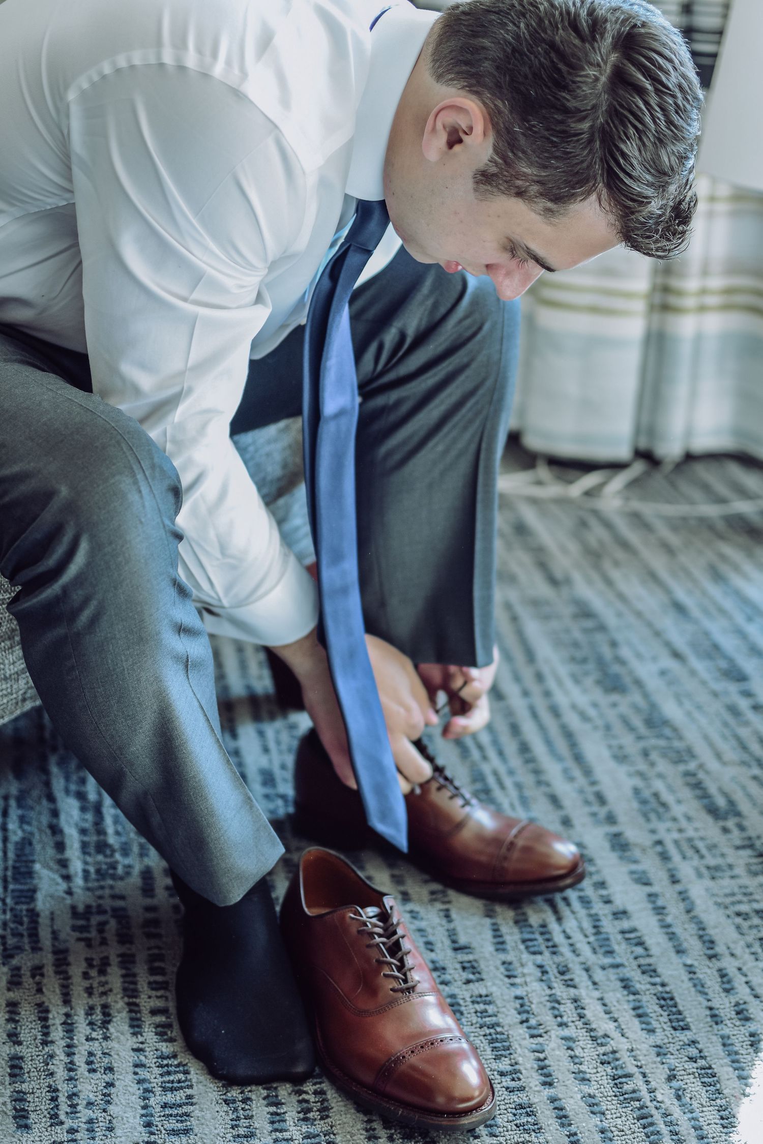 Person in business attire with blue tie tying brown leather dress shoes against a patterned carpet background.