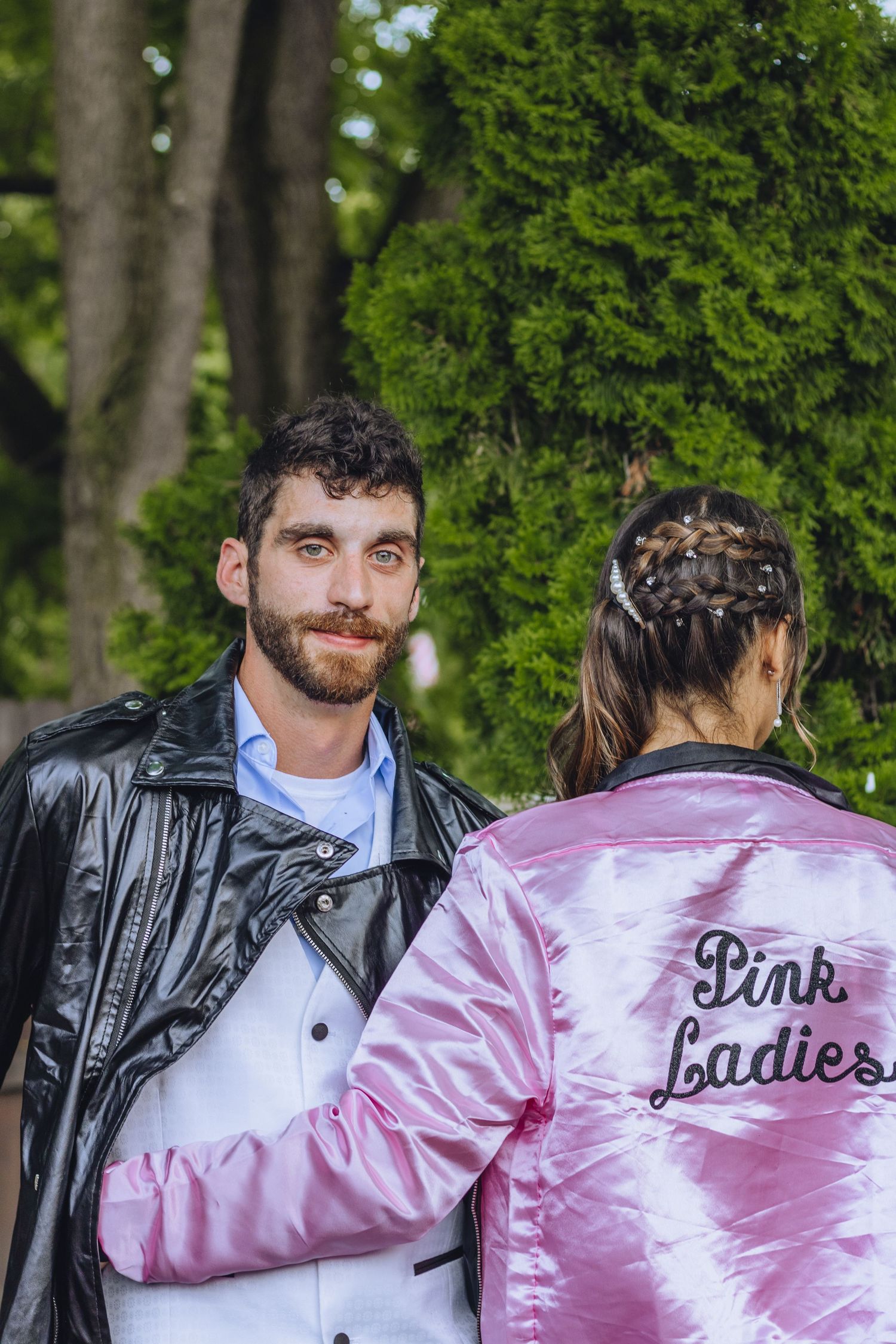Two people wearing jackets stand together outdoors, with one wearing a pink satin Pink Ladies jacket.