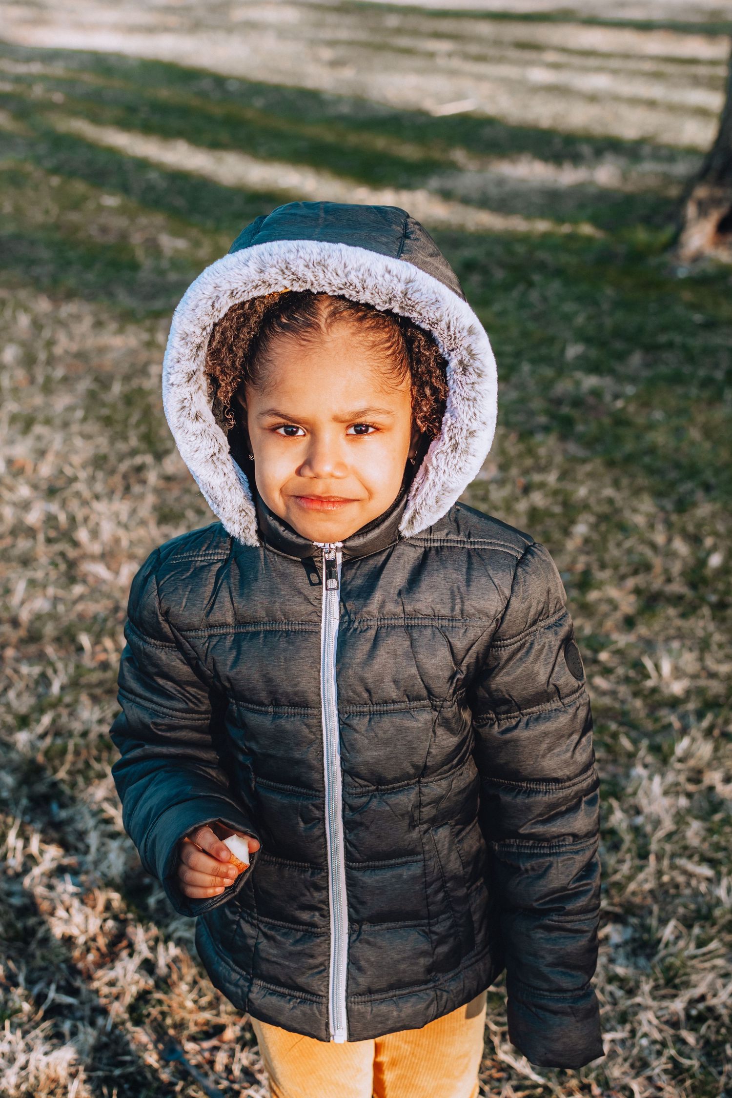 A child in a puffy winter jacket with fur-lined hood smiles at the camera outdoors.
