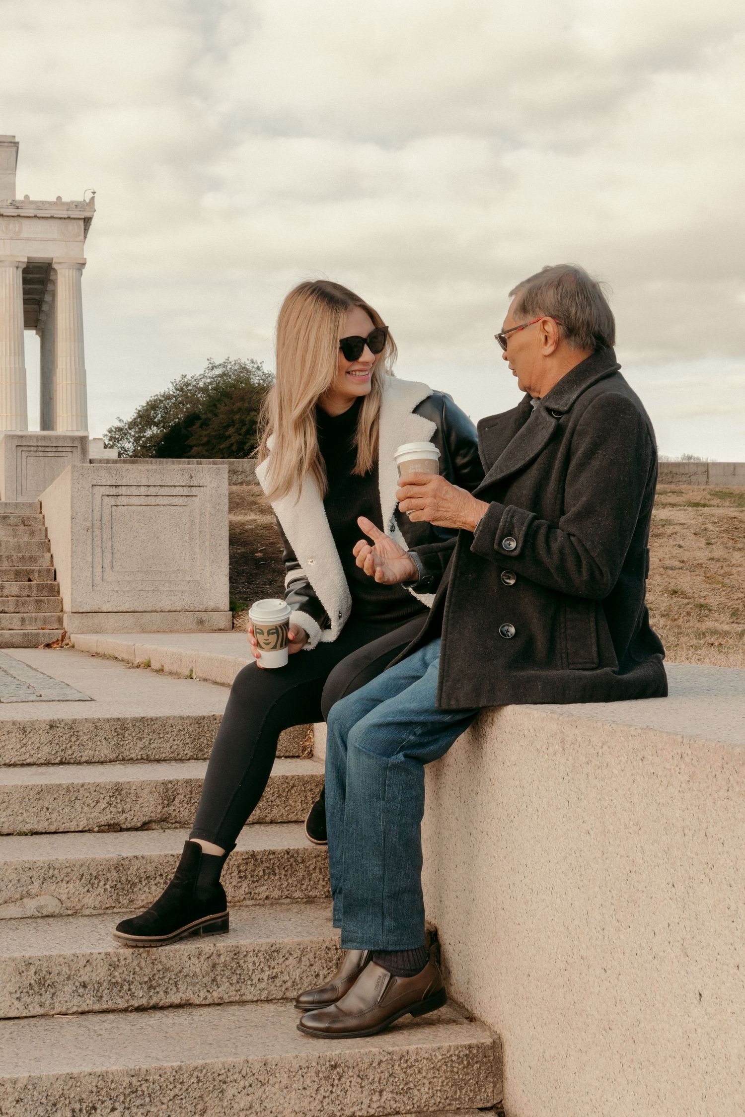 Two people enjoying coffee while sitting on stone steps near a classical structure.