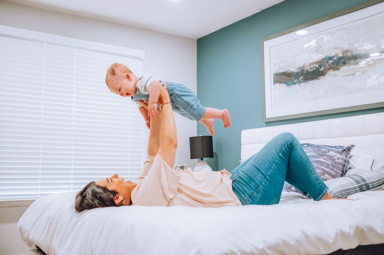 Playful moment as parent lifts baby wearing blue overalls while lying on white bed.