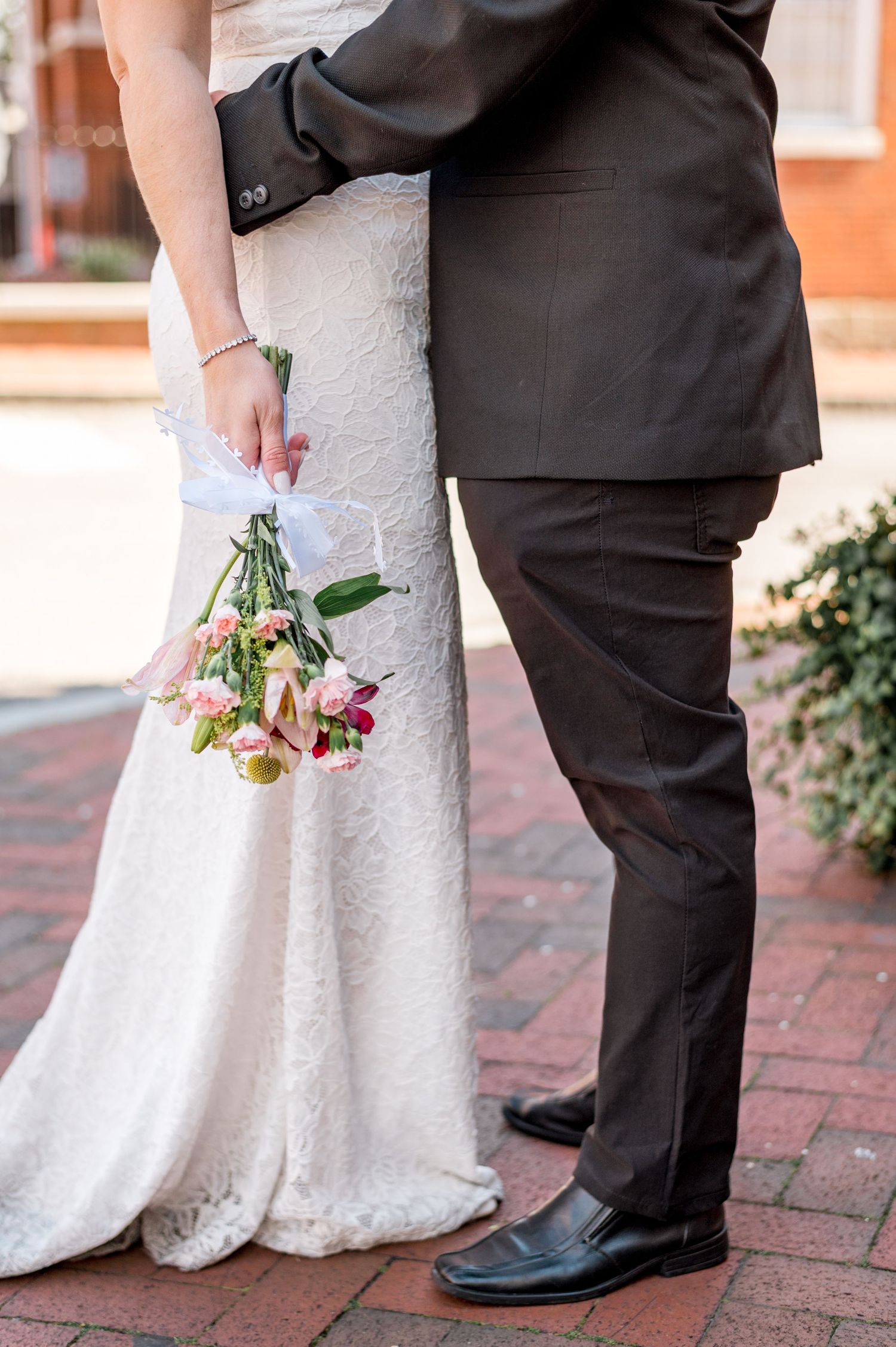 Detail shot of wedding couple with white lace dress and small bouquet on brick pathway.
