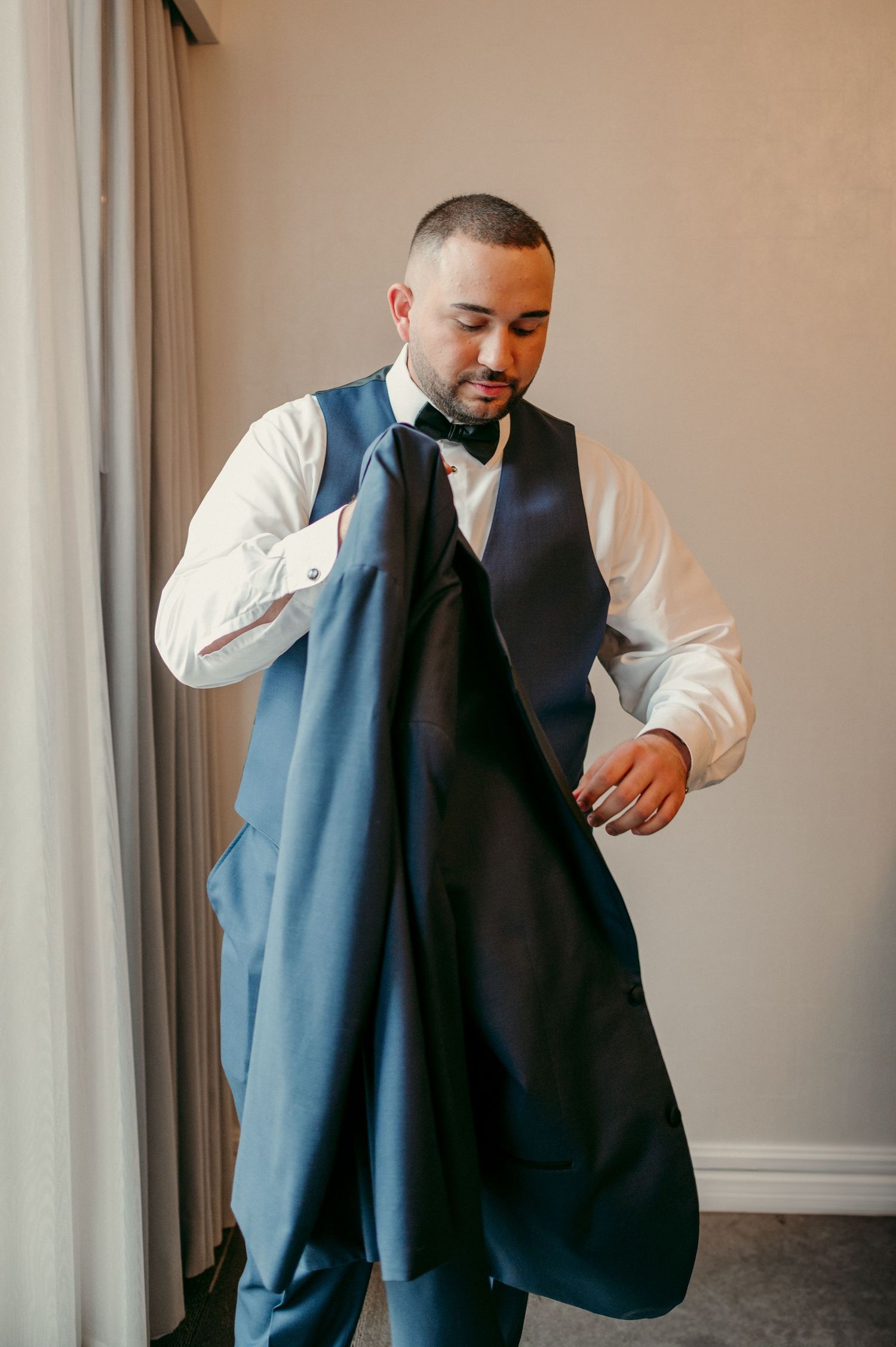 Someone getting dressed in formal attire with a navy blue vest, white dress shirt and bow tie while standing by a window.