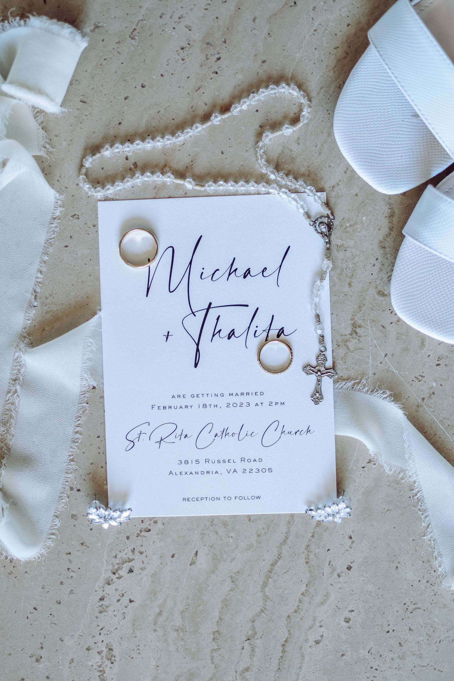 Wedding invitation with a gold ring and white ribbons arranged on a textured beige surface with white decorative details.