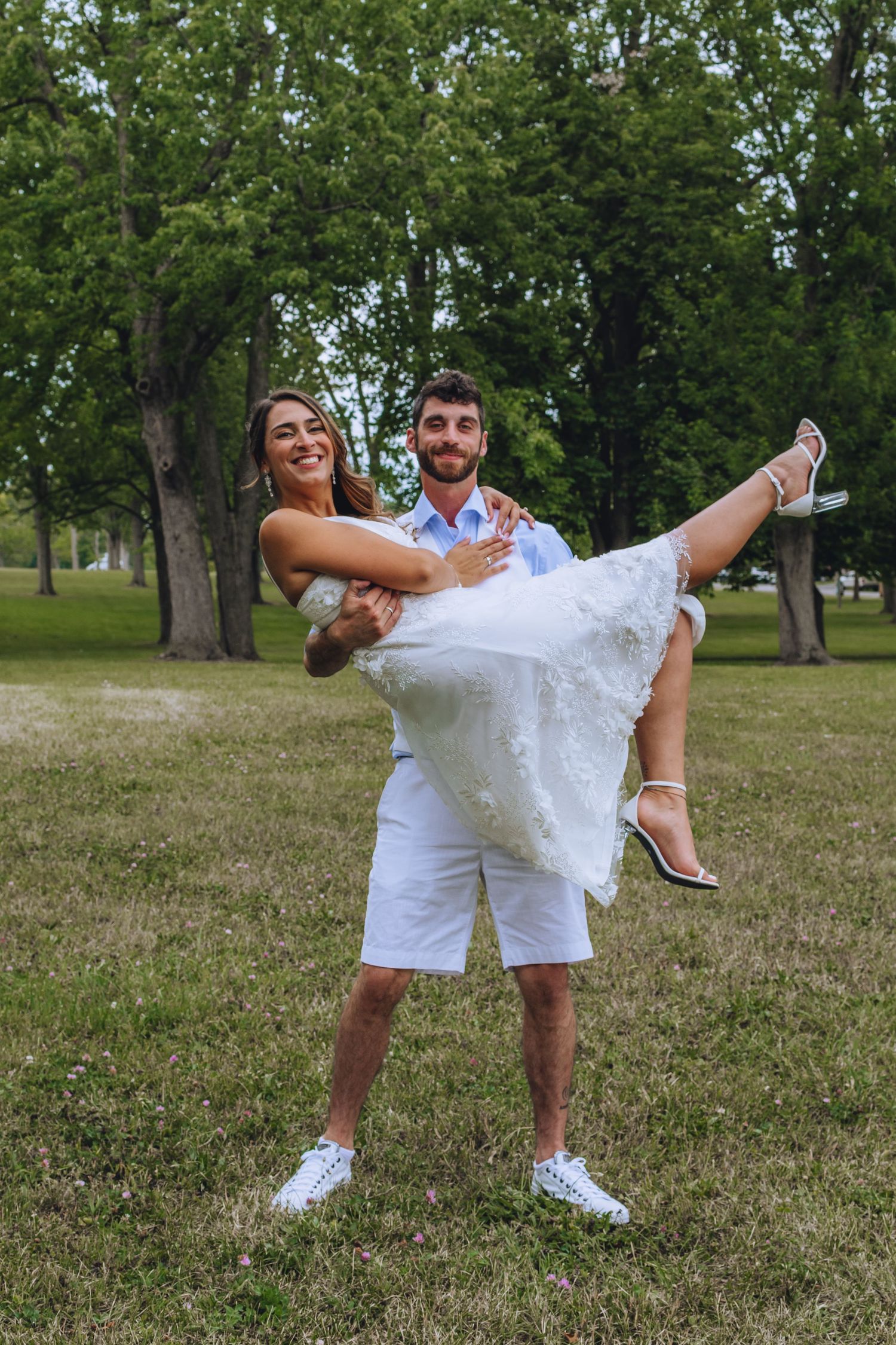 Two people wearing white outfits playfully pose together on grassy lawn in outdoor summer setting.