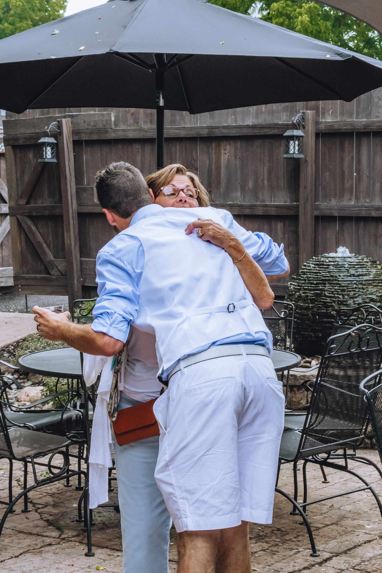 Two people embracing on an outdoor patio with a black umbrella and wrought iron furniture in the background.
