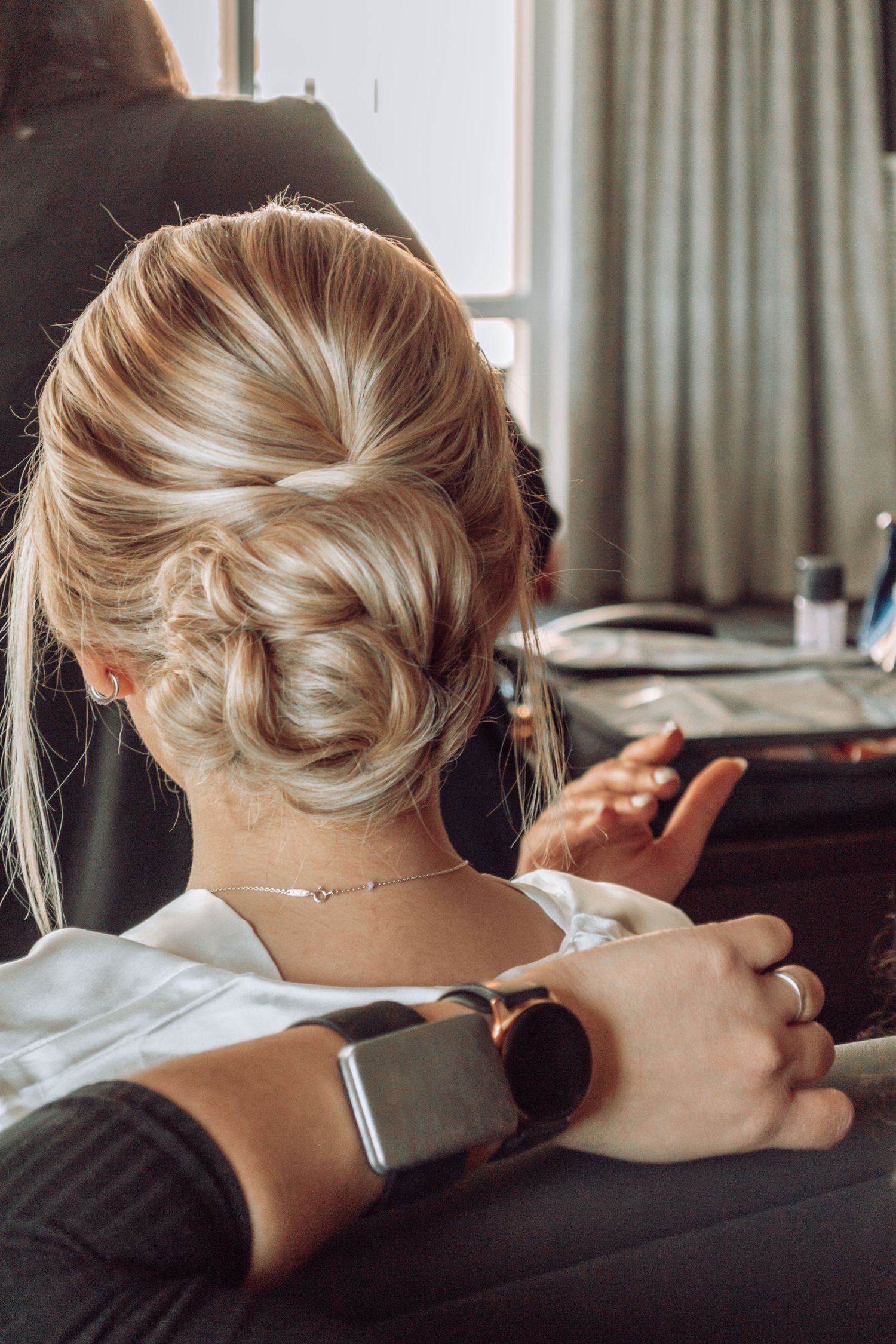 Elegant blonde updo hairstyle with twisted bun shown from behind as person checks their smartwatch in natural light.