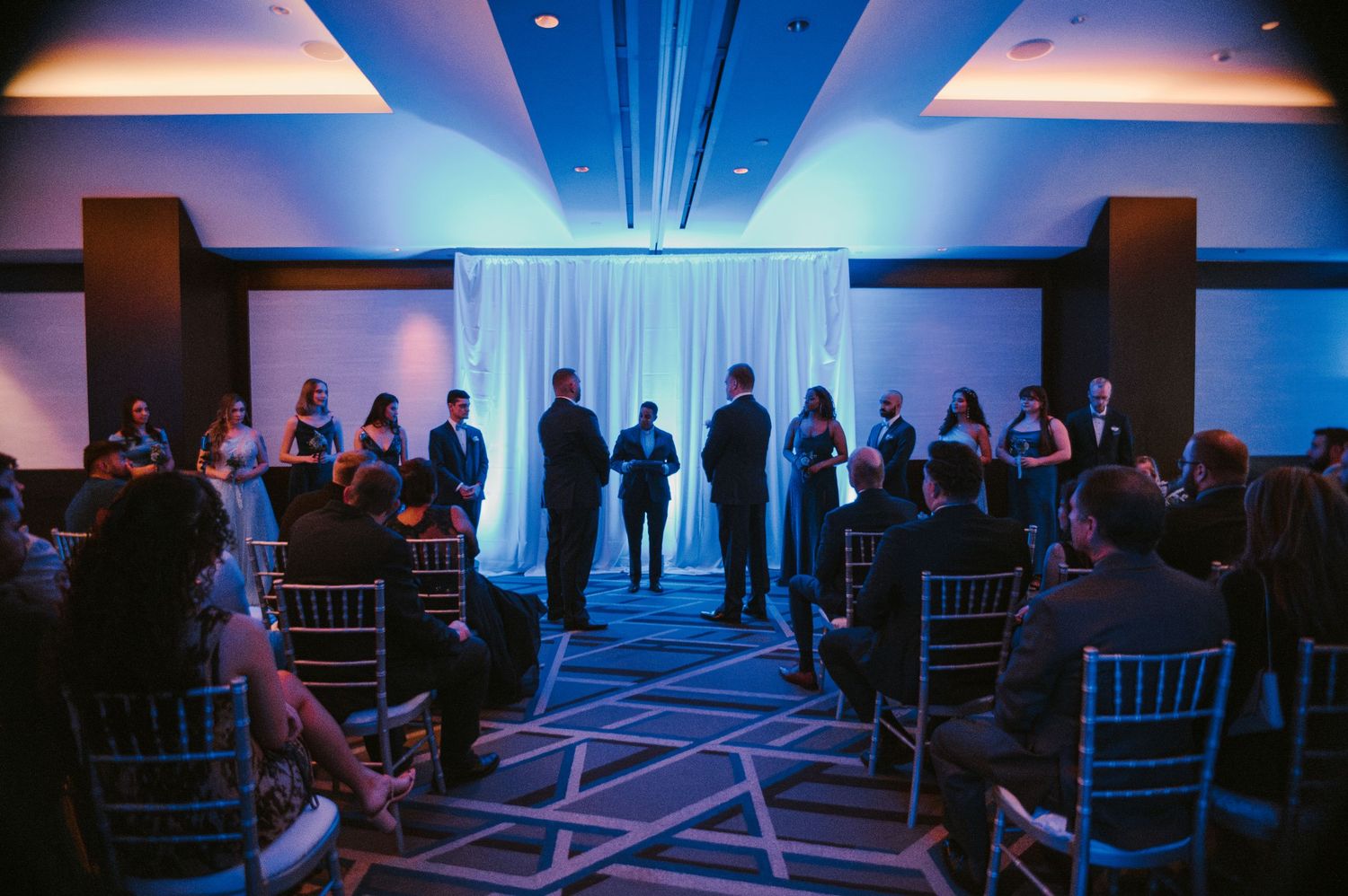 People attend an elegant ballroom event illuminated by blue lighting with a mix of standing and seated guests.
