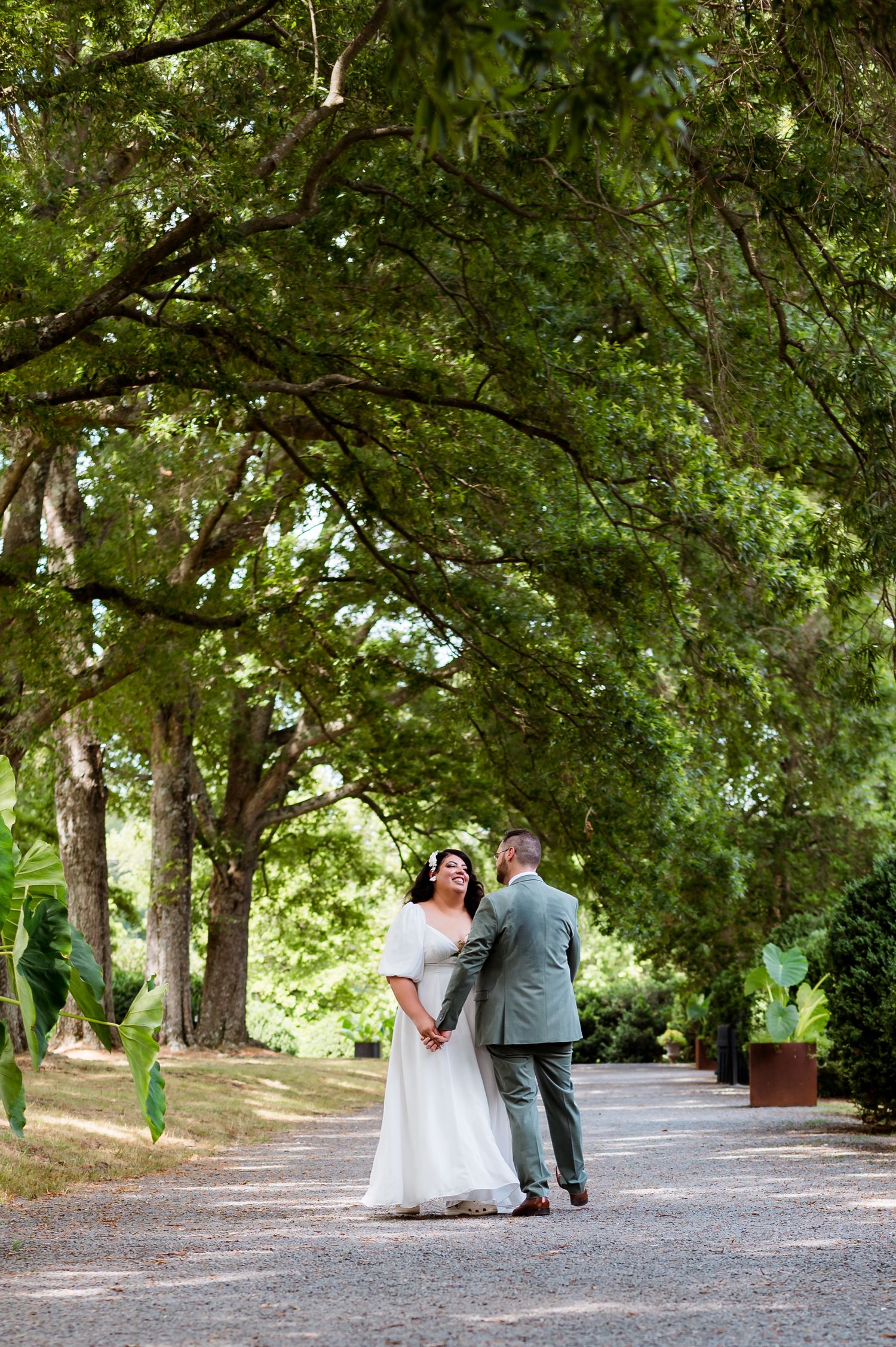 A couple stands together on a tree-lined gravel path during their outdoor wedding photoshoot.