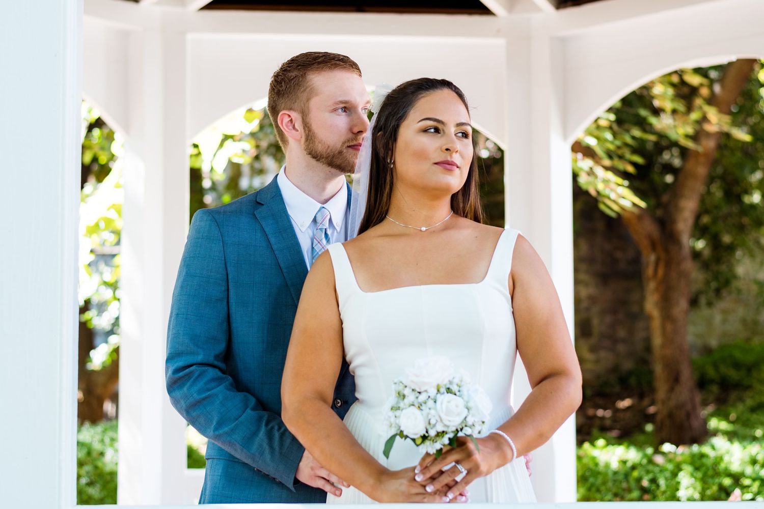 Elegant wedding portrait of couple standing in white gazebo with bride holding bouquet.