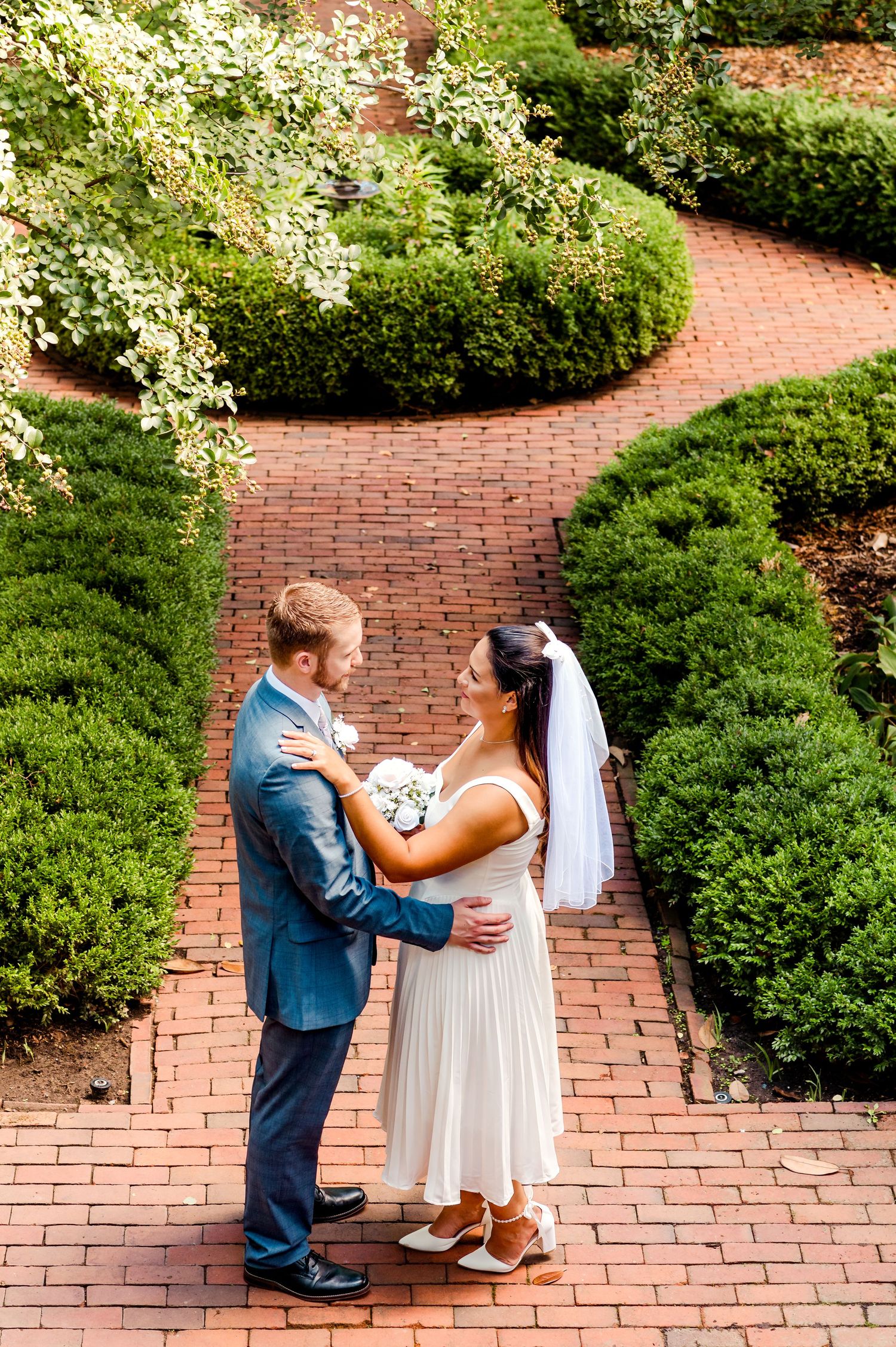 Couple shares romantic moment on brick garden path surrounded by flowering bushes.