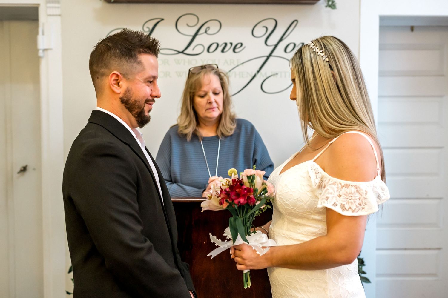 A bride in a lace off-shoulder dress shares an intimate moment with a groom in black suit during their indoor ceremony.
