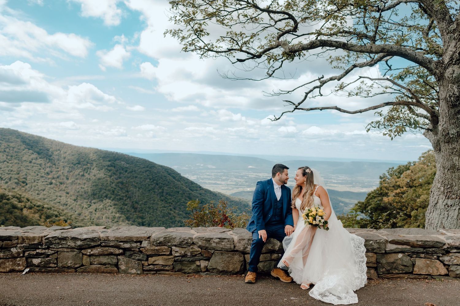 A happy couple in wedding attire embraces on a scenic mountain overlook with panoramic valley views in the background.