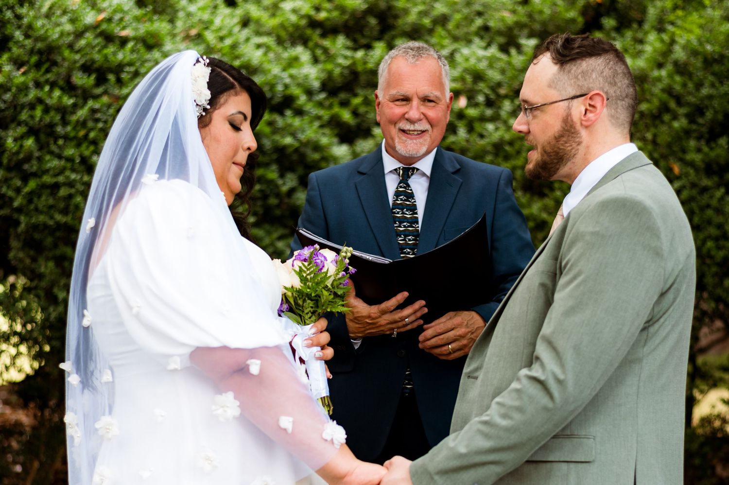 Wedding ceremony in a garden setting with an officiant leading the service between a bride in white and partner in sage green.