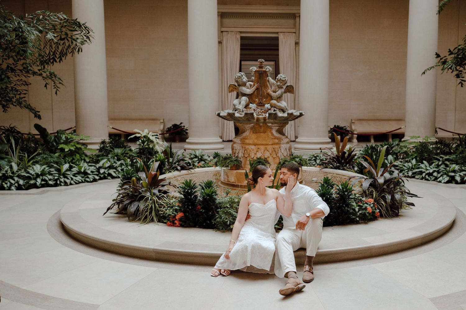 Couple sitting together near ornate stone fountain surrounded by tropical plants in elegant courtyard.