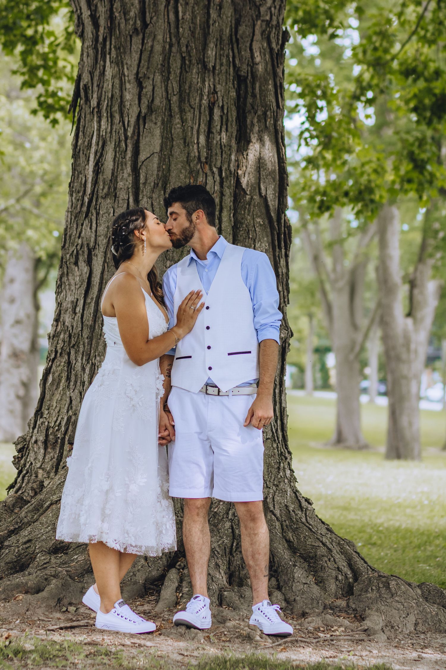 A romantic couple dressed in white outfits share tender moments against a tree trunk in a sunlit park setting.
