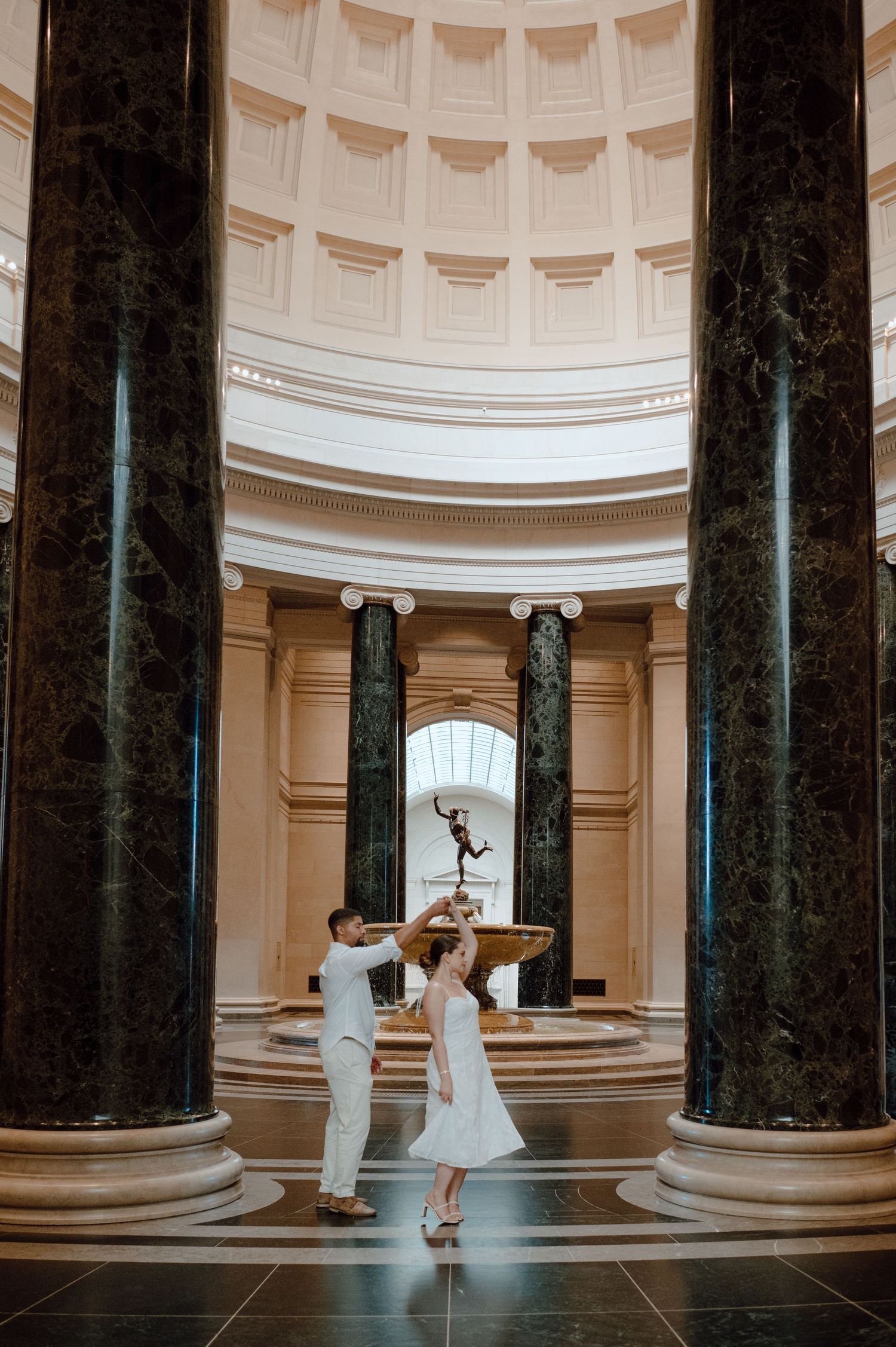 A couple dancing in an elegant domed room with marble columns and a classical statue centerpiece.