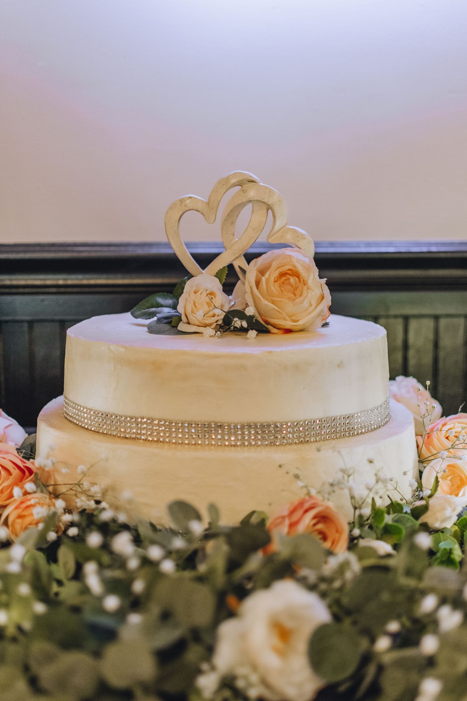 Classic white wedding cake with rhinestone ribbon and golden heart topper decorated with peach roses and greenery.