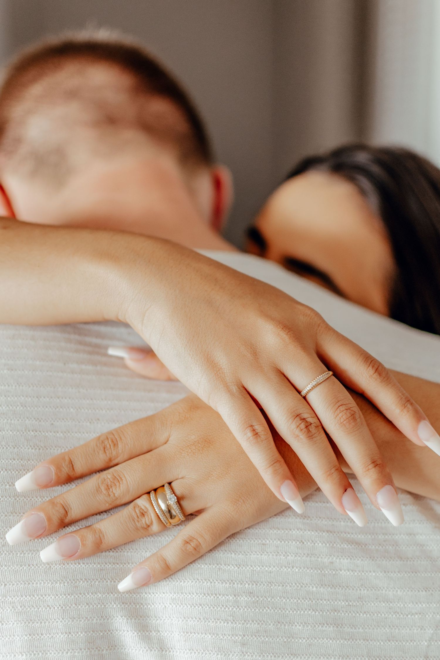 Close-up shot of hands wearing elegant wedding rings and French manicured nails resting on white fabric.