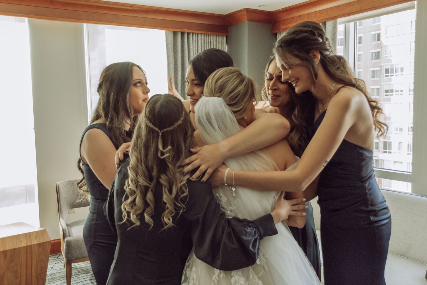 A group of wedding attendants in navy dresses share an emotional embrace by a window during pre-ceremony preparations.