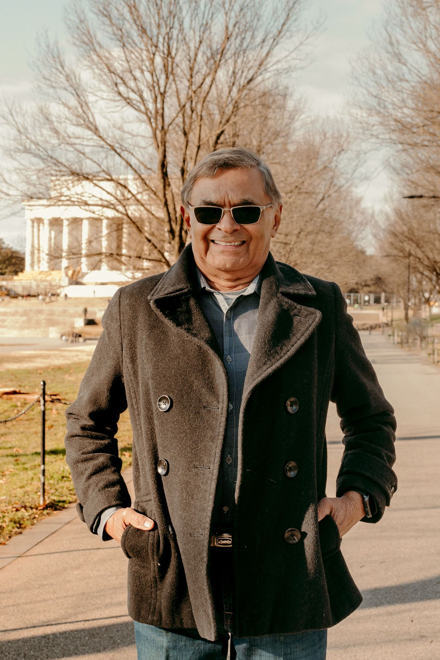 Person in peacoat and sunglasses stands confidently on pathway with bare trees in background.
