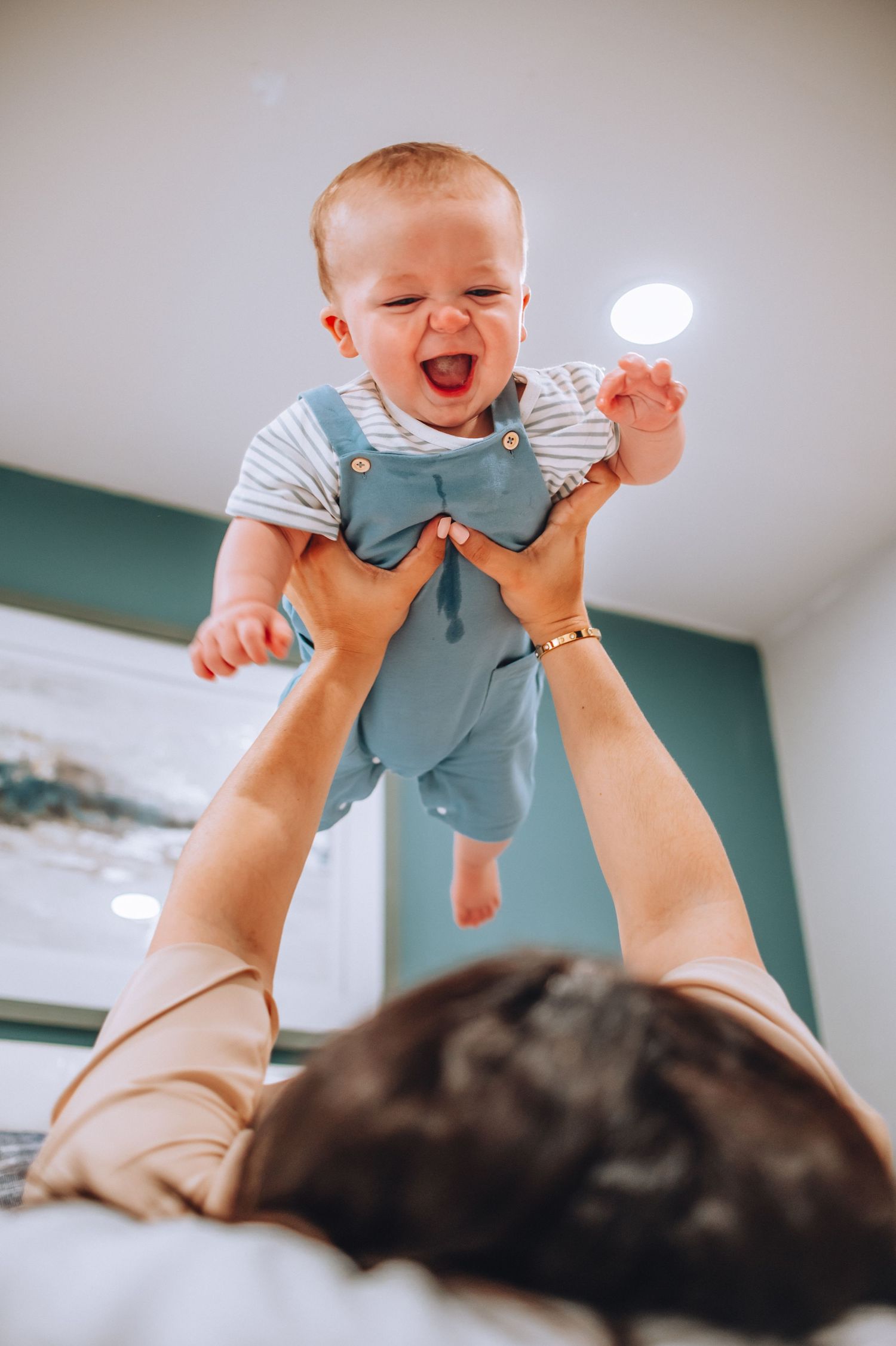 Baby wearing blue overalls is lifted high in the air during playtime on bed.