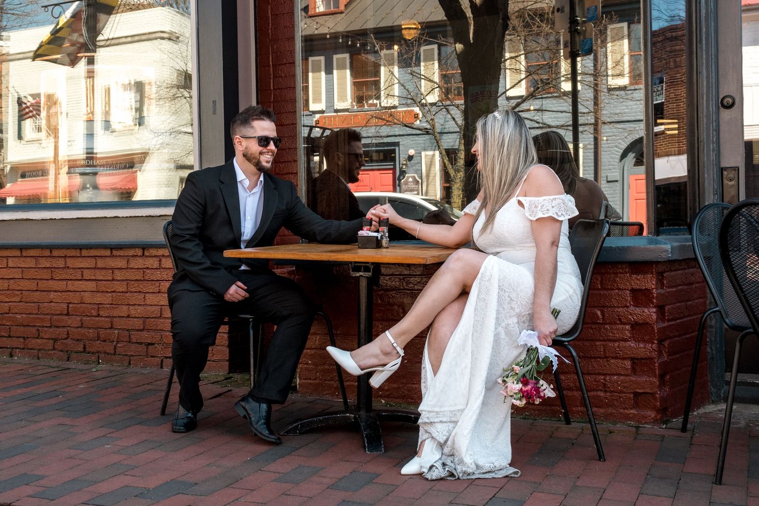 Bride and groom share drinks at an outdoor cafe table in an urban setting.