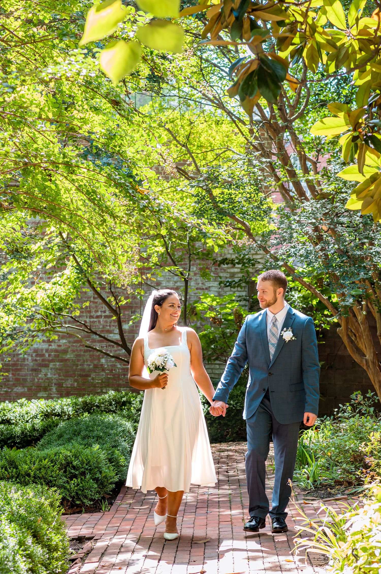 A couple in wedding attire walks hand in hand along a brick path surrounded by lush greenery and trees.