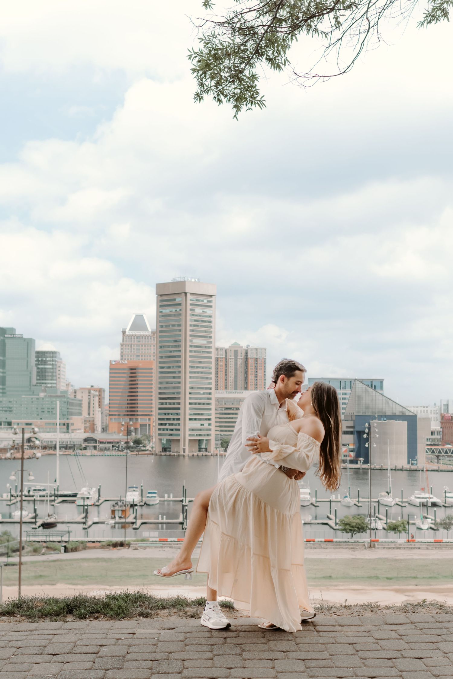 A romantic couple embracing against the Baltimore Inner Harbor skyline and marina on a cloudy day.
