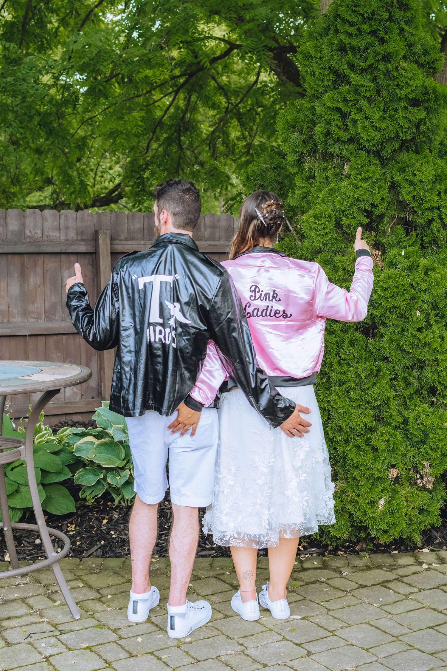 Two people wearing matching retro style jackets, bomber style pink and black, pose together in a garden setting.