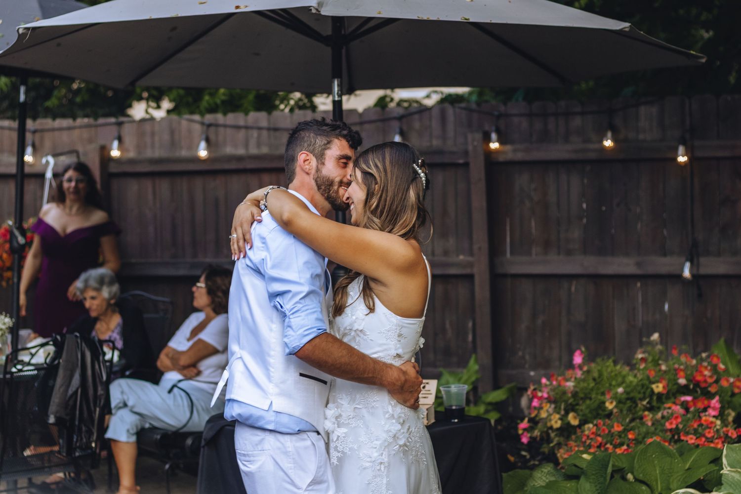 A couple in formal attire dances closely together at an outdoor wedding reception with string lights and flowers.