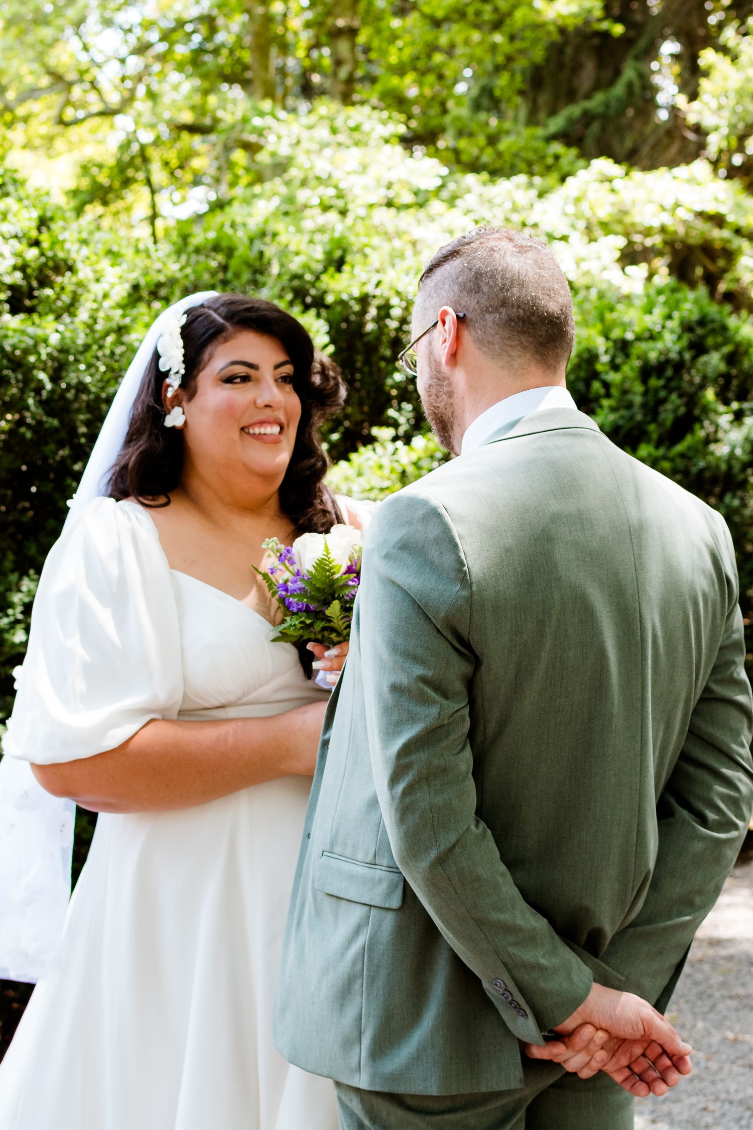 Bride in flowing white dress and veil shares a tender moment with groom in sage green suit in a garden setting.