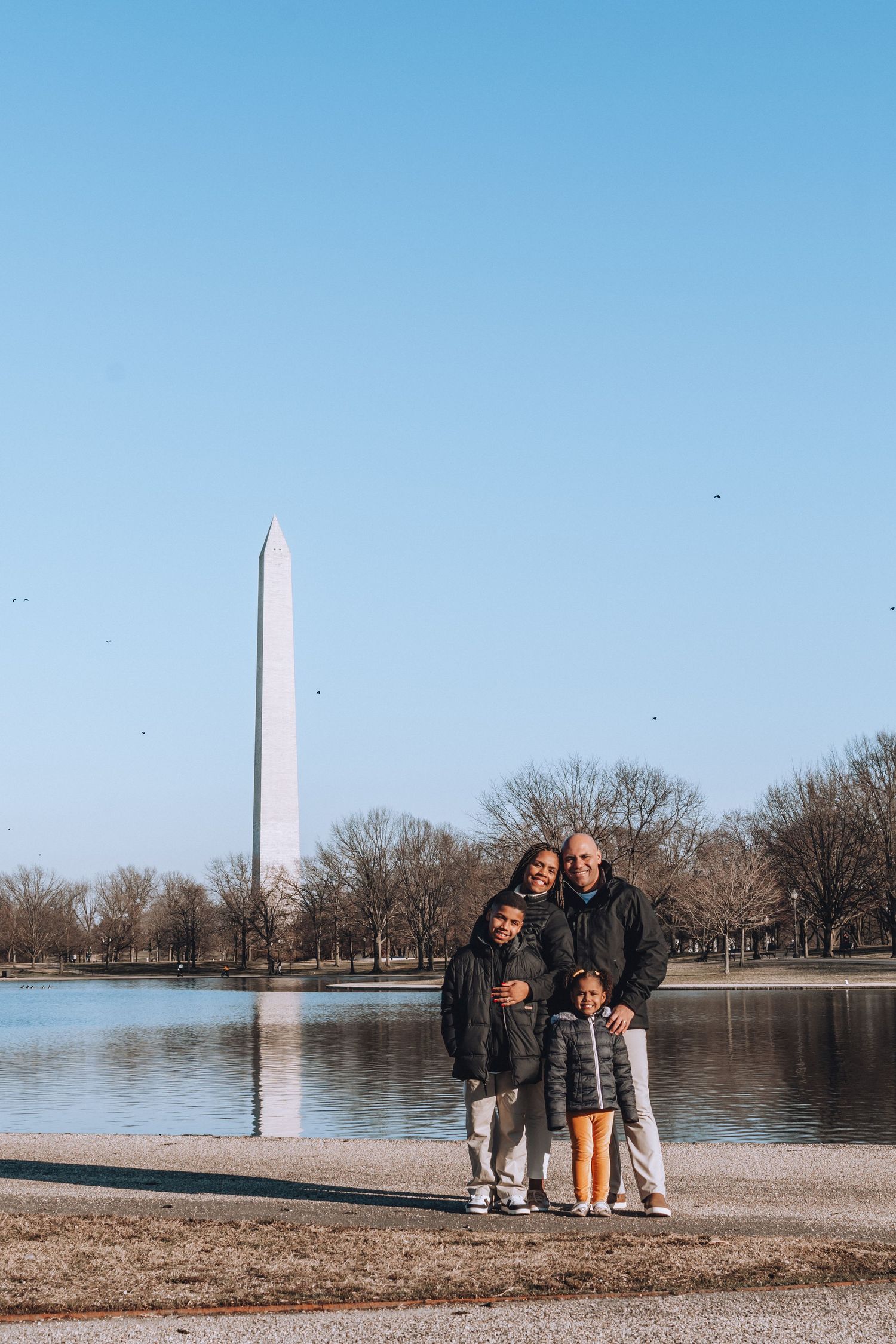 Family standing by reflecting pool with Washington Monument towering in background on clear winter day.