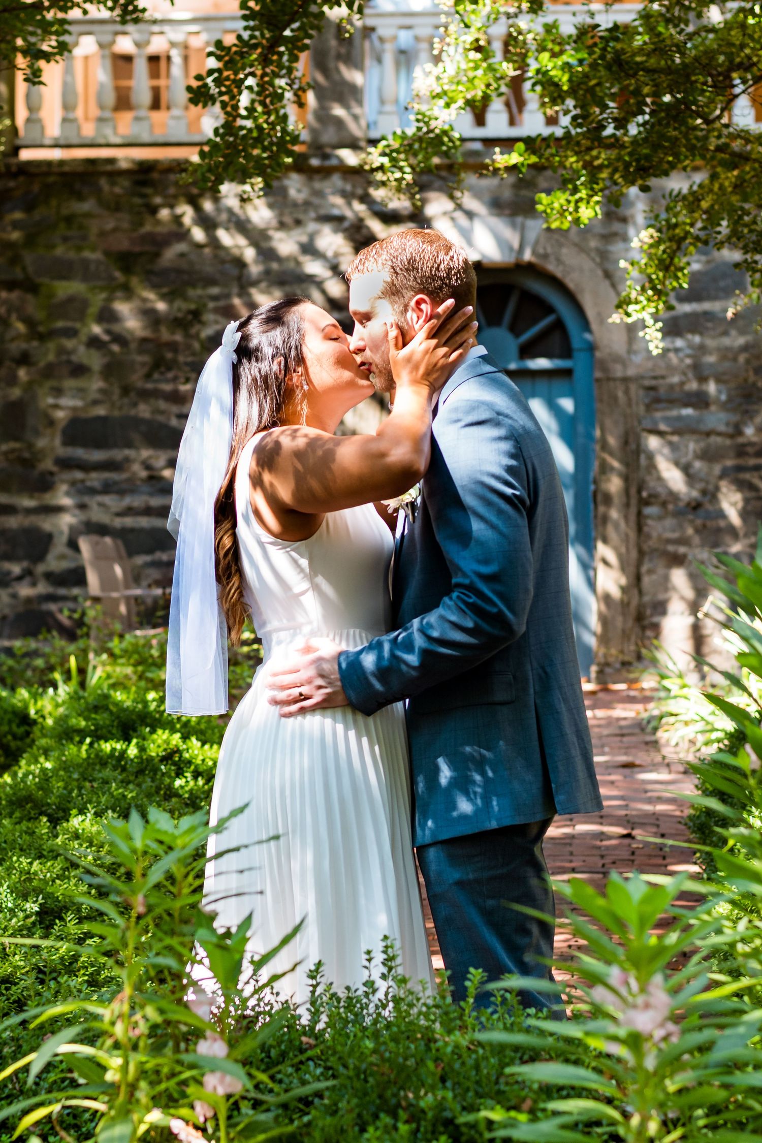 Romantic wedding kiss in garden setting with blue door and stone wall backdrop.