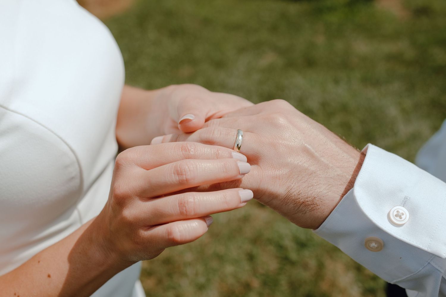 Close-up view of hands exchanging wedding rings during an outdoor ceremony with grass in the background.