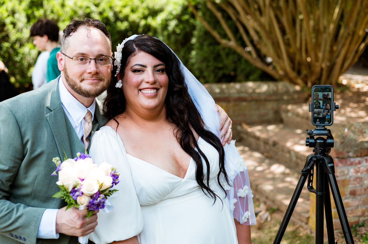 A couple in wedding attire poses outdoors with a tripod and bouquet of purple flowers at a garden venue.