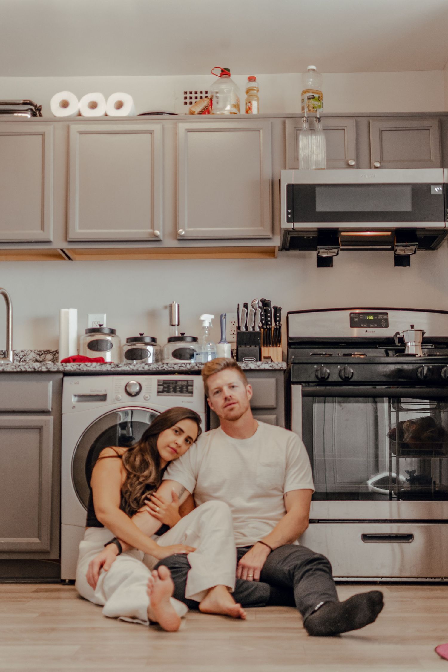 Cozy couple sits on kitchen floor with gray cabinets, appliances, and household items stored above.