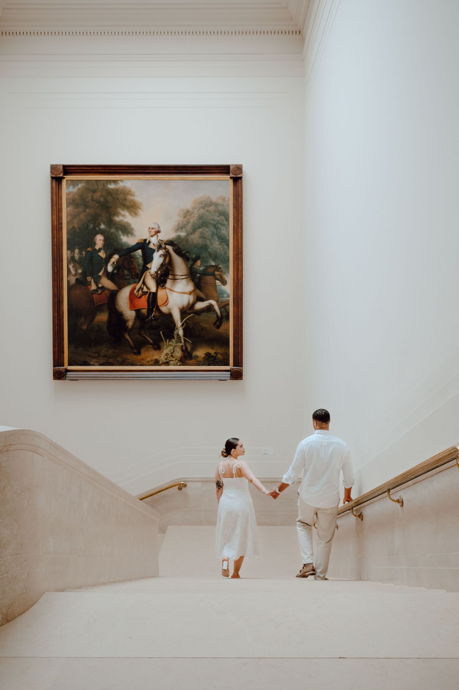 A couple in wedding attire walks up elegant stairs past classical paintings in a museum or gallery setting.