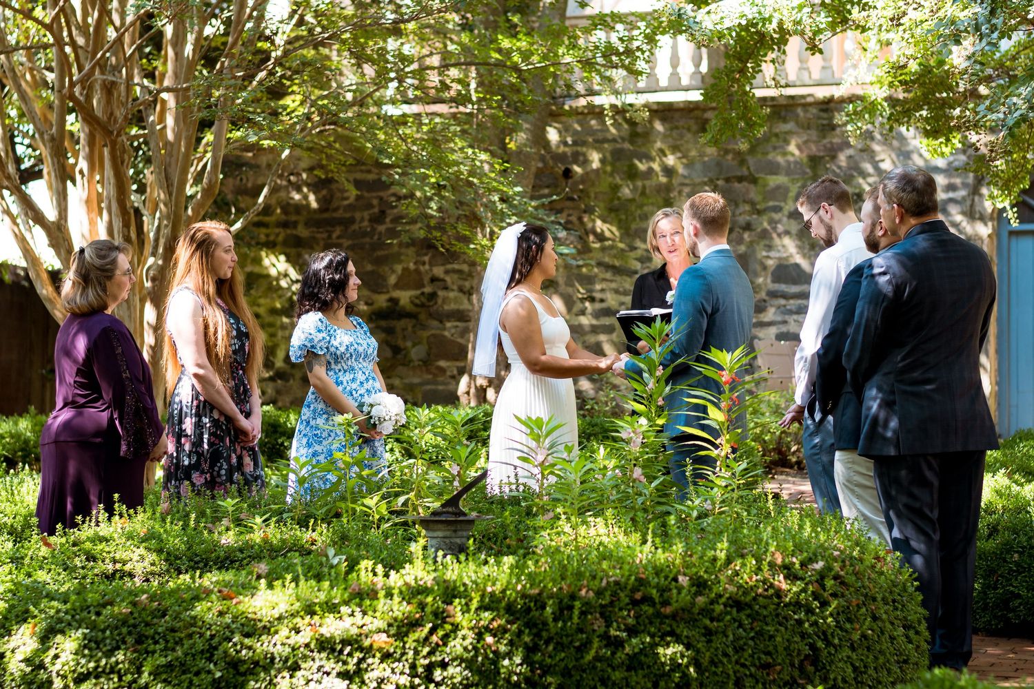 Group of wedding guests enjoying an outdoor garden reception on a sunny summer day amidst lush greenery and stone walls.
