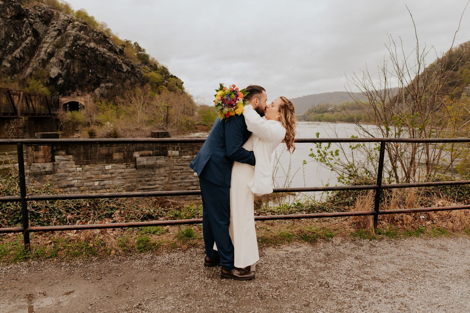 A couple in wedding attire embrace on a rustic overlook with rocky cliffs and mountains in the background.