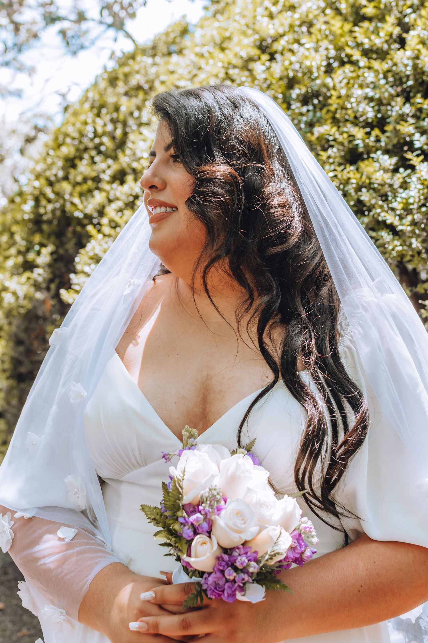 Joyful bride in white dress holds purple and white bouquet while smiling under her veil against sunny garden backdrop.