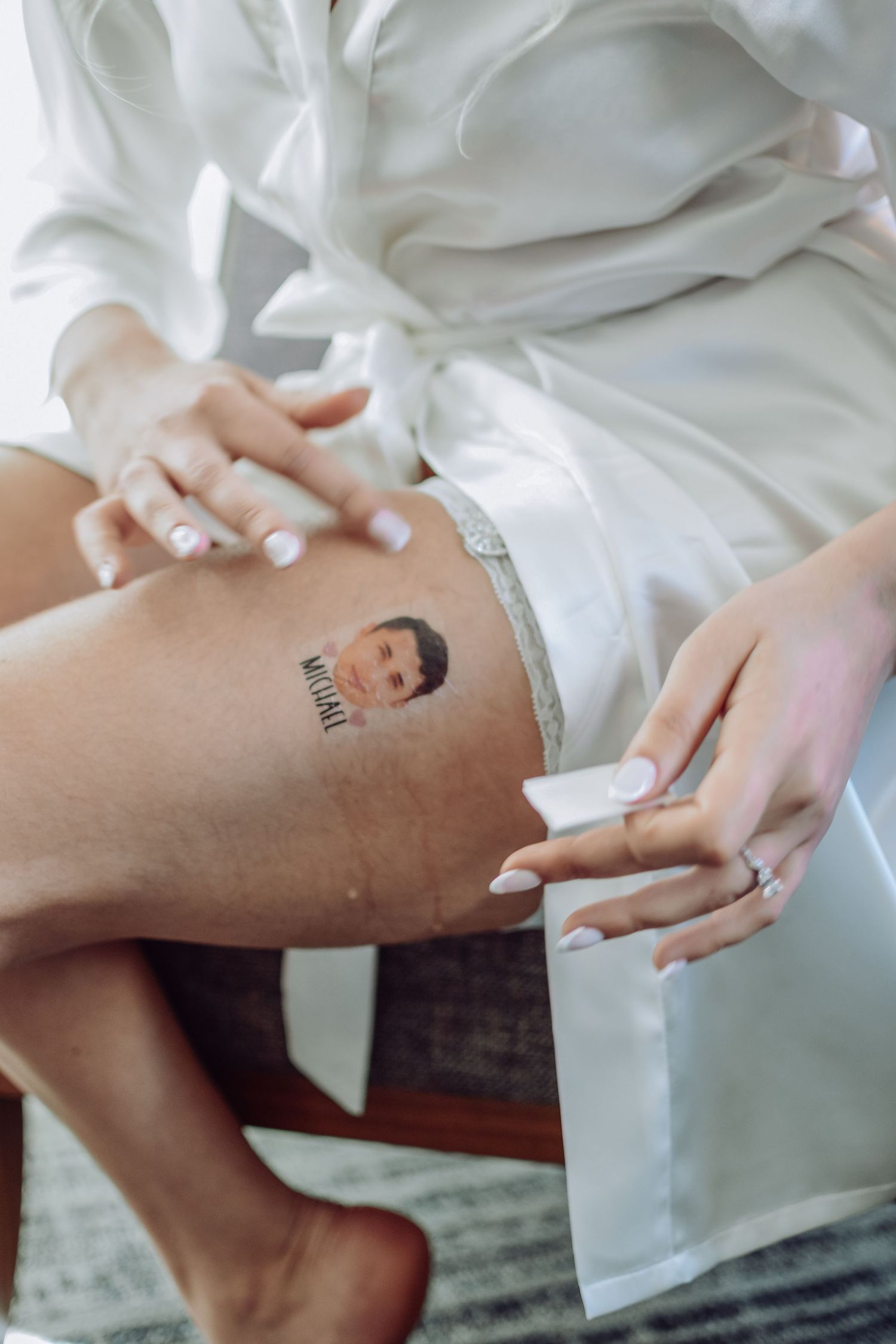 Person in white robe showing tattoo on thigh with decorated nails and a ring on their hand.