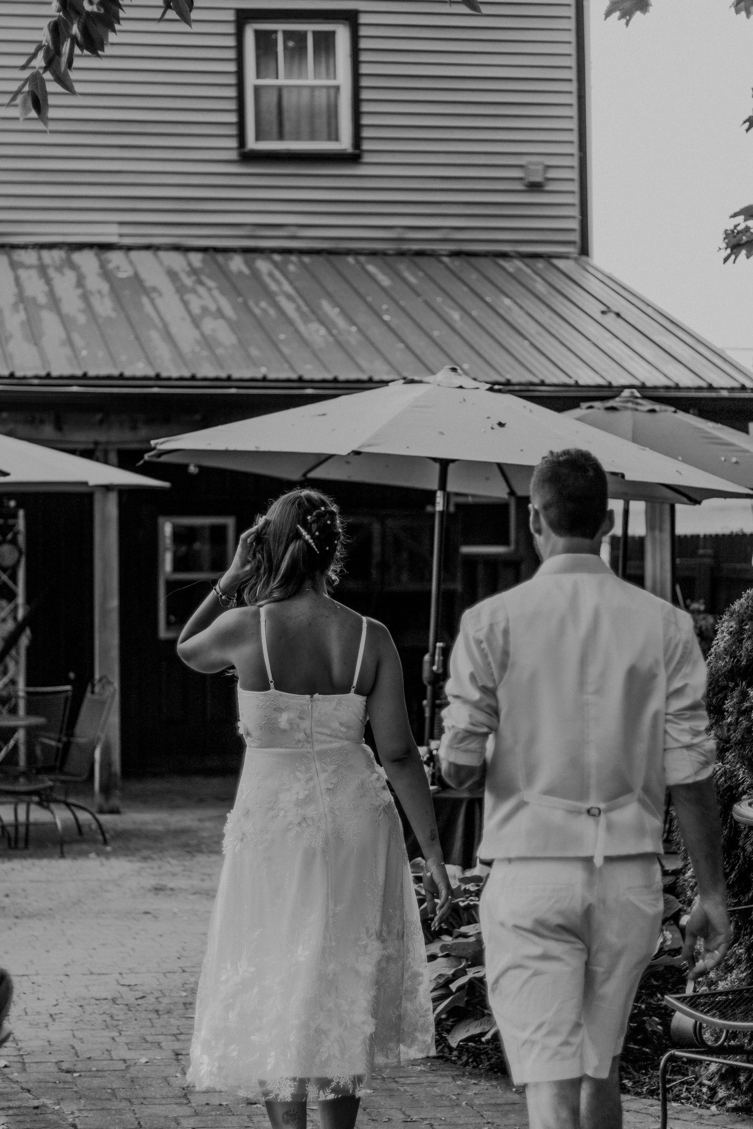 People carrying umbrellas walk on a dirt path in front of wooden buildings in a black and white vintage scene.