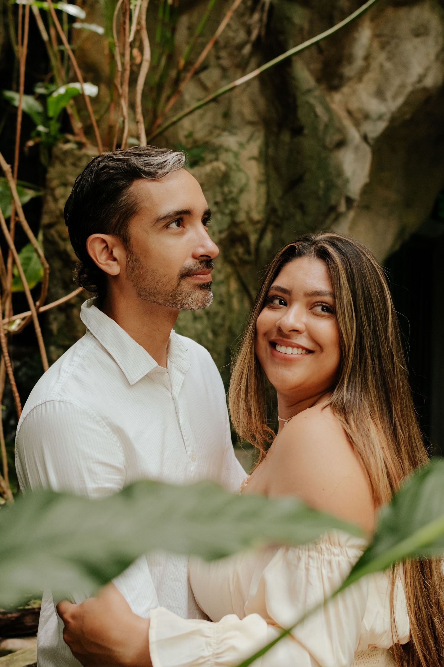Loving couple in white outfits pose together among tropical foliage and stone walls.