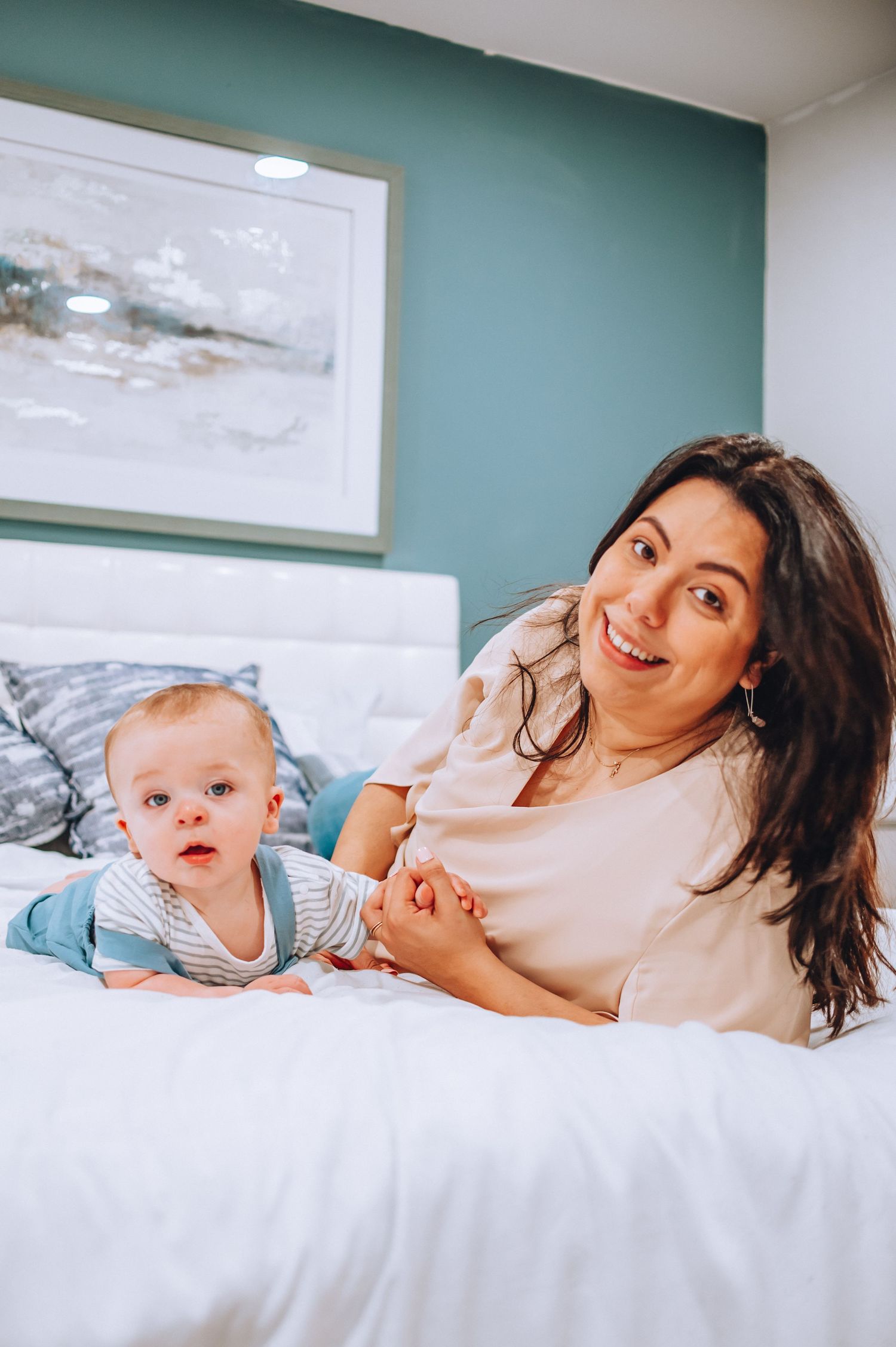 Mother and baby relaxing together on white bedding with teal accent wall and artwork.